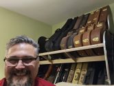 the face of a man, wearing glasses, smiling in front of shelves filled with guitar cases