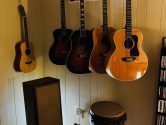5 acoustic guitars hanging from the wall, in the corner of a room, above a speaker and drum.
