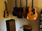 5 acoustic guitars hanging from the wall, in the corner of a room, above a speaker and drum.