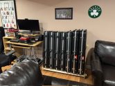 7 black guitar cases standing up in a wood rack, next to couches and a desk with a computer