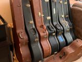brown and black acoustic guitar cases standing up in a  wood rack, next to one on the floor, and a couch