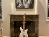 white stratocaster in a guitar stand in front of a fireplace with a Beatles poster above