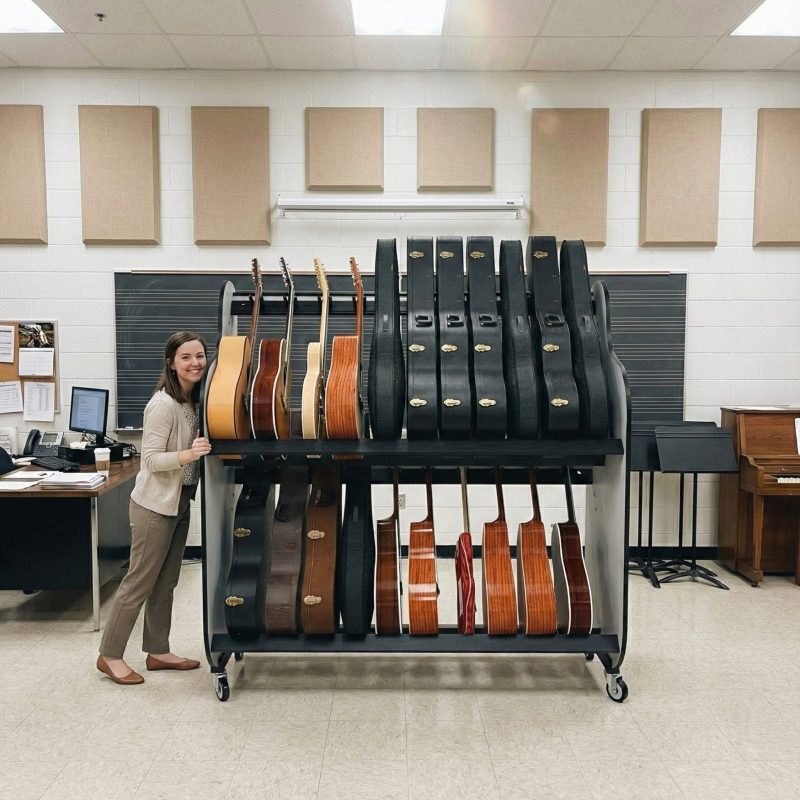 A female teacher pushing a two-tiered guitar shelf rack in a music classroom setting