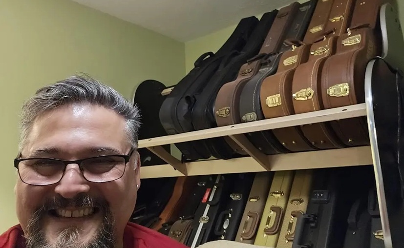 man smiling while sitting in front of shelves that are filled with guitar cases
